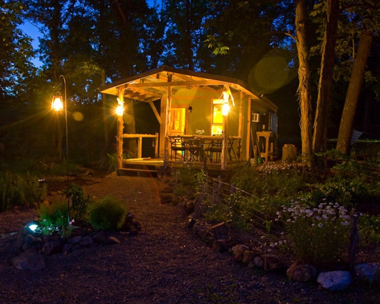 A tiny house in the woods in the evening. Several lanterns light the porch and a flower garden in front of the cabin.