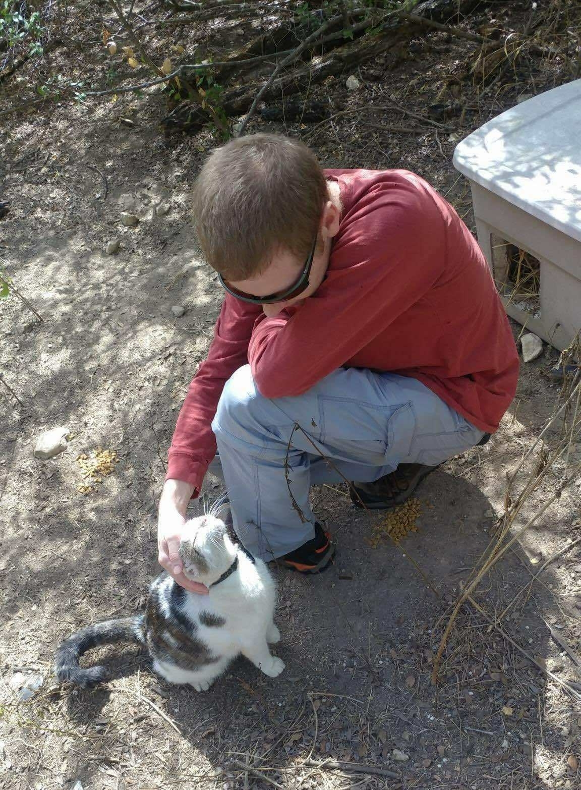 Photo of short haired man in red shirt and blue pants kneeling down and petting a white and gray cat