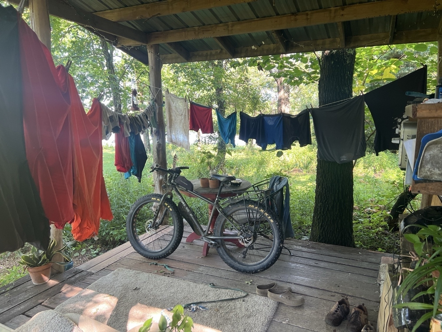 Two laundry lines strung across a wood porch deck with a variety of colorful clothing hanging to dry. Also on the porch, a fat-tire bike leans against a table. There are several shoes visible on the deck and a woodland in the background.