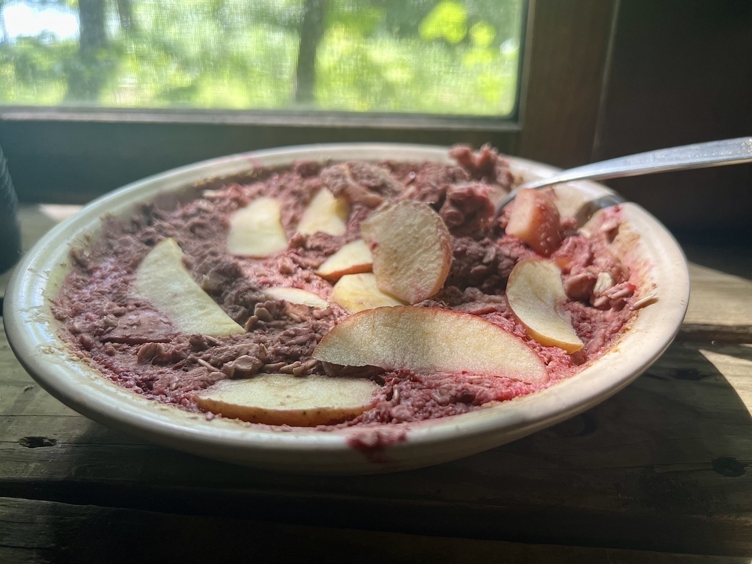 A yellow bowl containing baked oatmeal in front of a window. The oatmeal is mixed with blended blackberries so it's purplish pink with apple wedges across the top