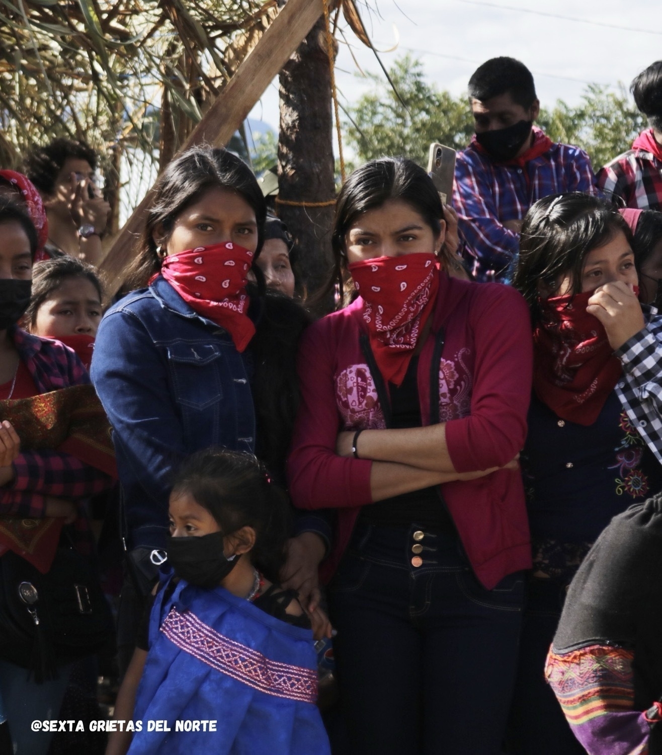 Three indigenous women in the foreground of an image, a young girl in front of them are wearing bandanas on their faces. Around and behind them are other indigenous people similarly masked