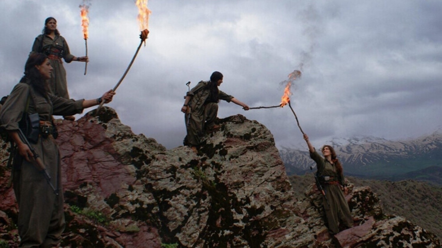 4 women in dark green uniforms standing on a rock peak with mountains in the background. The women each hold a lit torch with the fourth woman holding her torch up as it is being lit by another