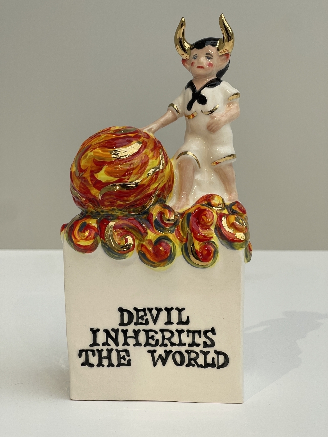 Ceramic figurine of small boy with horns touching a fiery, orange and gold orb with title “Devil Inherits The World”