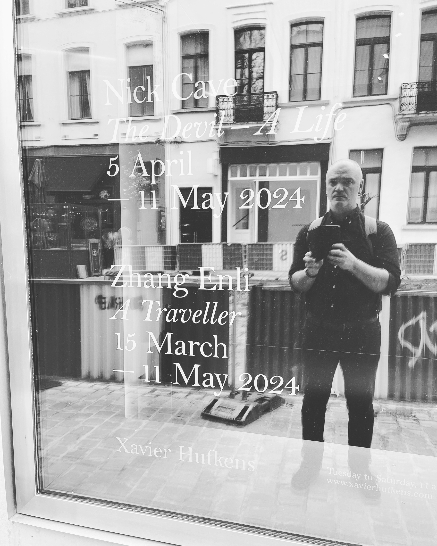 Benjamin Holsteen, reflected in the glass window of Xavier Hufkens Gallery in Brussels, Belgium. The window advertises the exhibition of Nick Cave’s _The Devil — A Life_ on display from 5 April - 11 May.