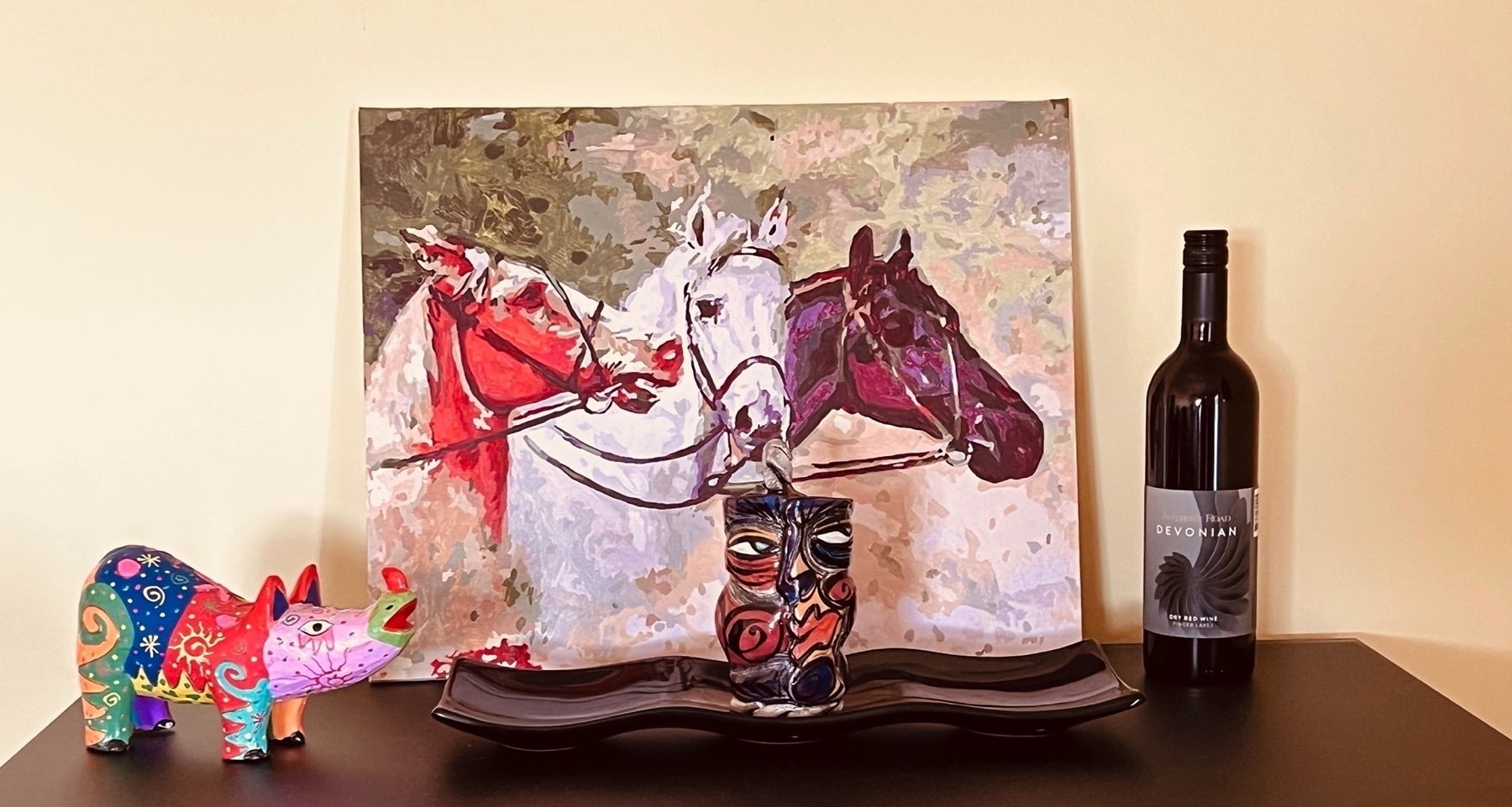 Five objects arranged on top of a liquor cabinet. From left to write: a brightly multi-colored wooden rhinoceros from Indonesia; a handmade mug with a cubist face painted on it (the mug is positioned so the handle is behind it); the mug is resting on black display tray; a bottle of red wine from the NYS Lake District; in the back, a painting of three horses.
