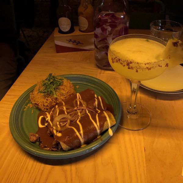  A plate with two enchiladas drizzled with vegan “cream”, and a cocktail with pineapple garnish.