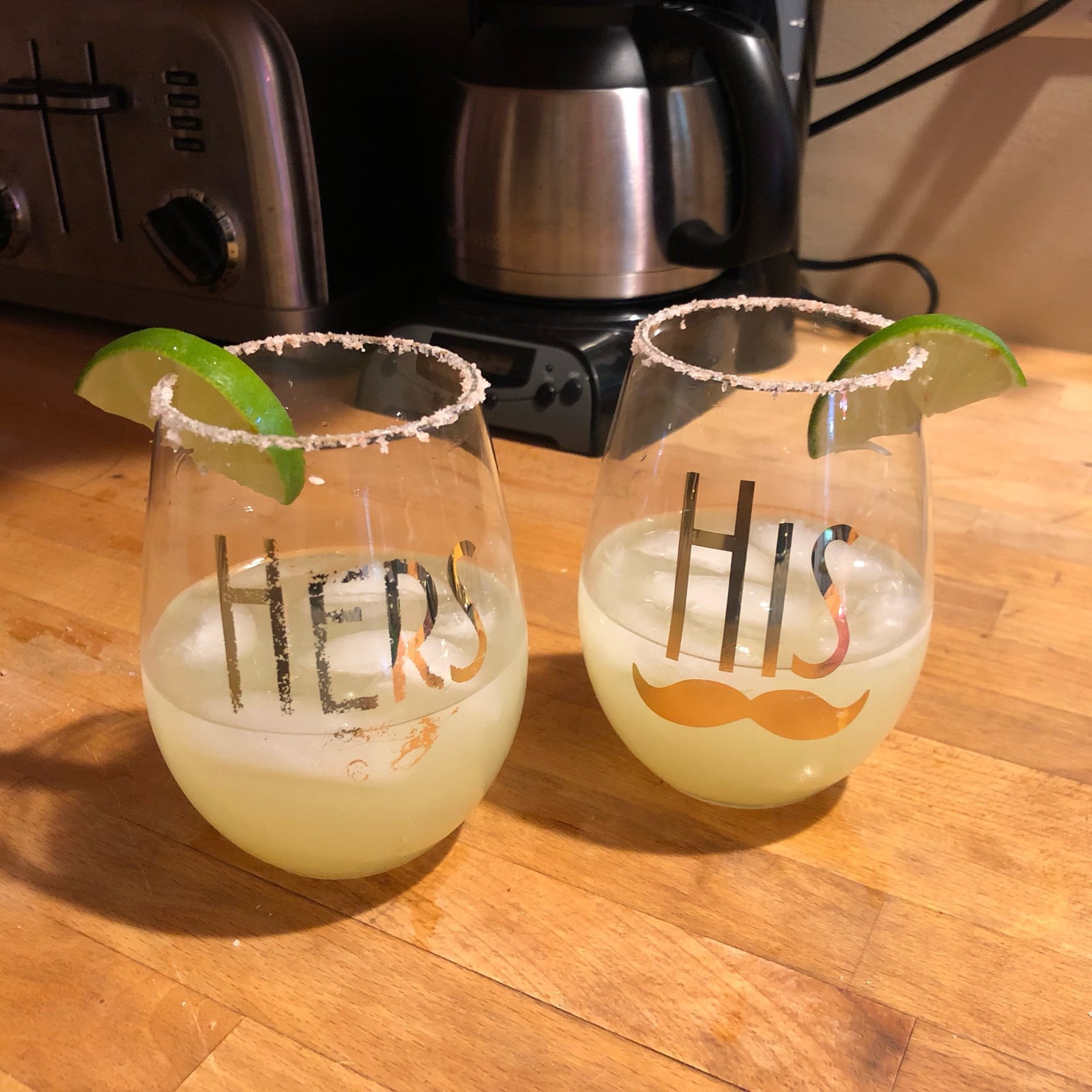 Stemless wine glasses, embossed with gold "His" and "Hers" decals, partly filled with ice and margarita cocktails.