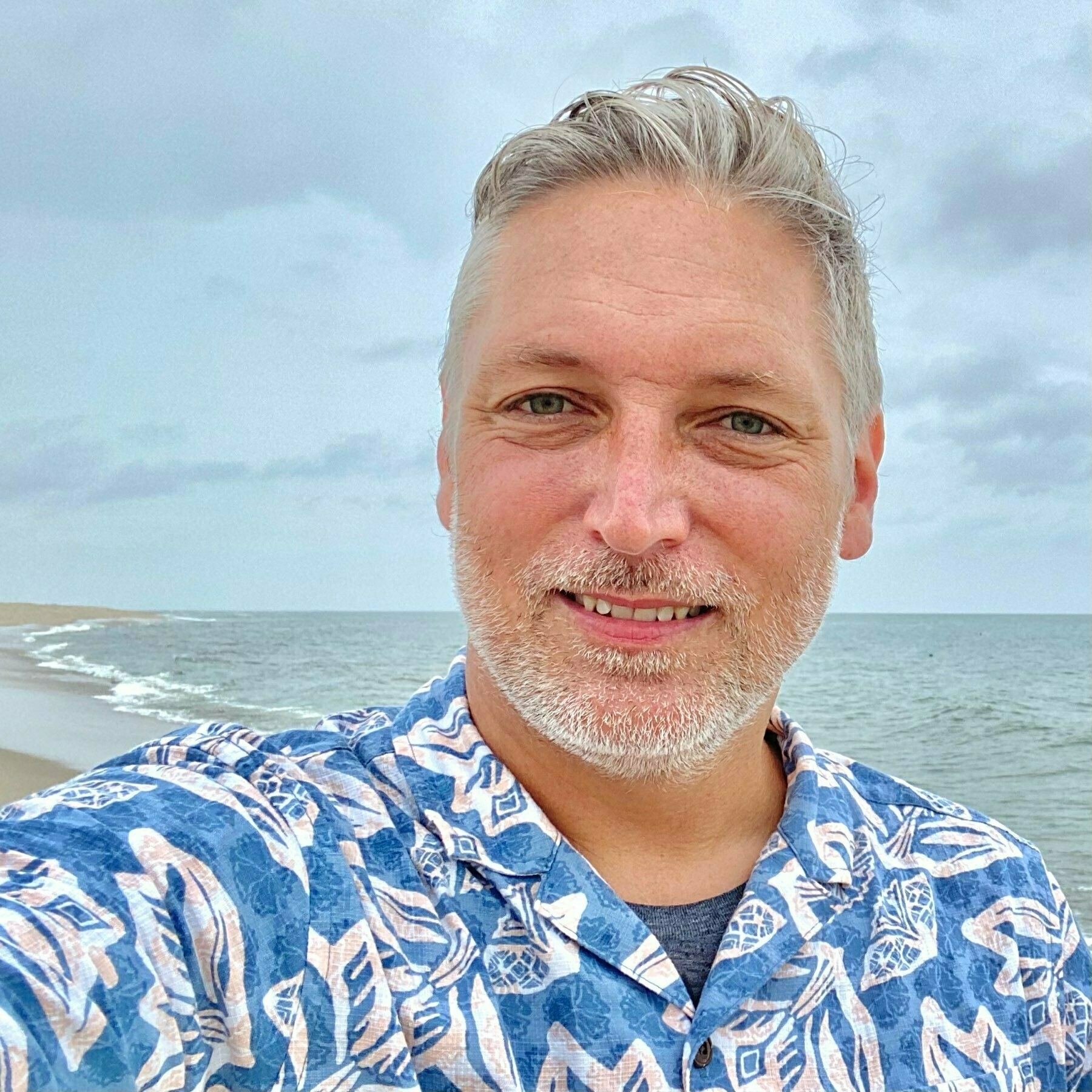 selfie against a backdrop of ocean and beach. i'm wearing a vaguely hawaiian style orint shirt, with grey stubble