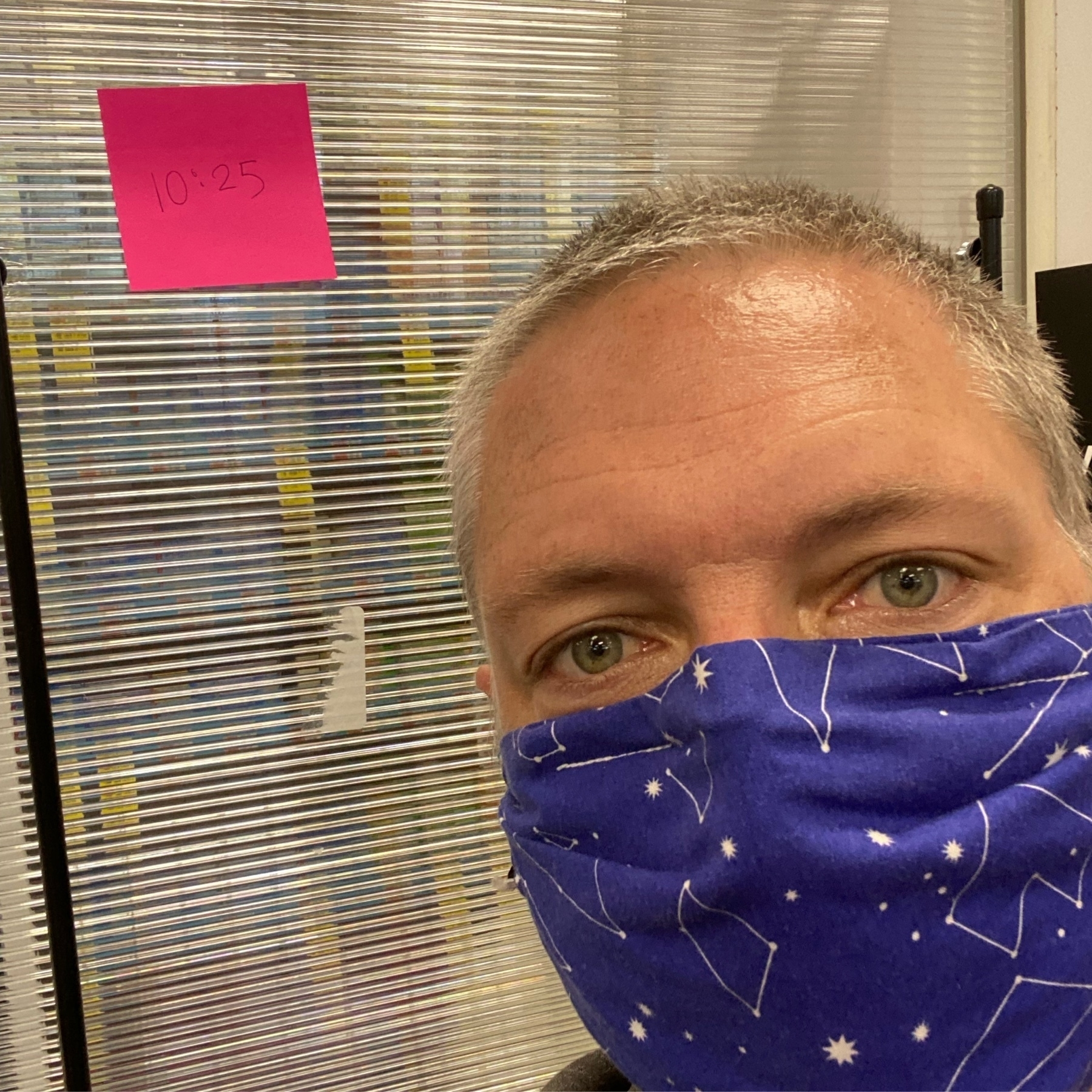 Self portrait wearing a constellation mask, beside a post-it note “10:25” in a pharmacy waiting area.