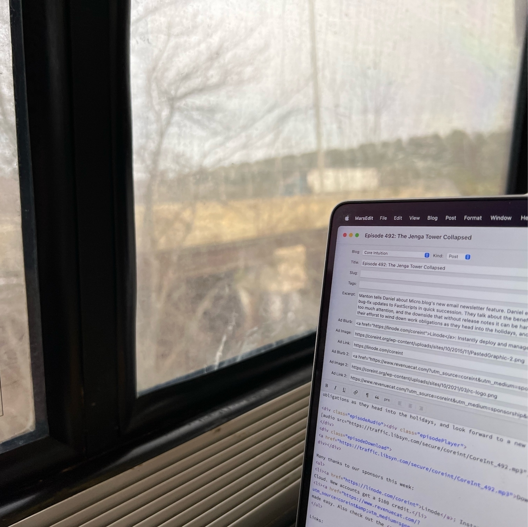 picture of a macbook pro opened to MarsEdit, editong show notes, with train window and landscape behind