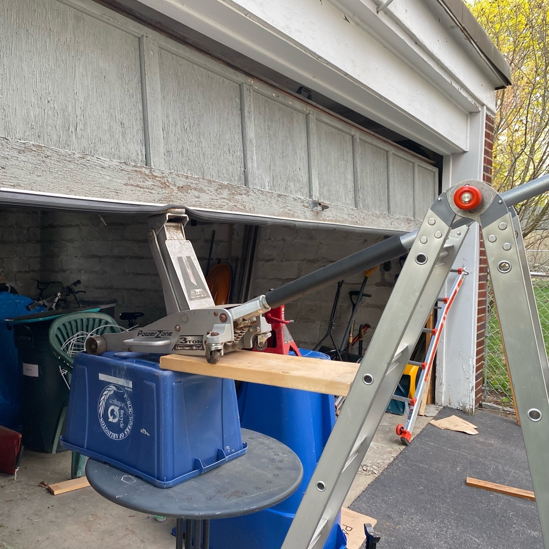 pictue of a garage door being held open by a contrived combination of table, recyling bin, and automotive floor jack