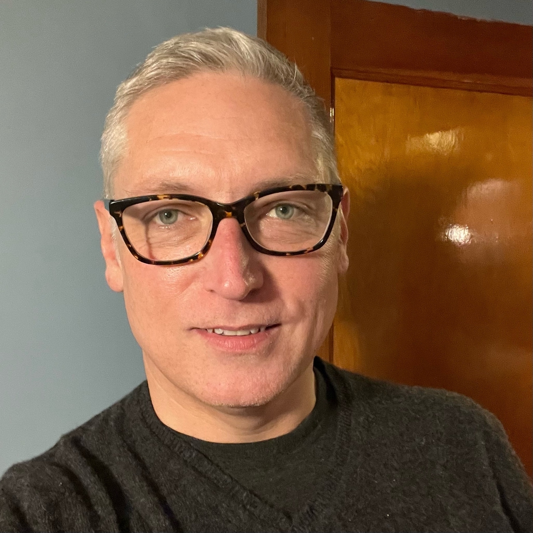 Selfie wearing a black sweater and glasses, looking into camera, shortly cut silver hair
