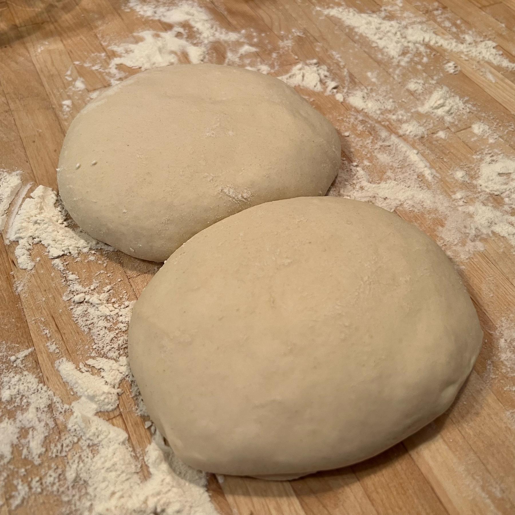 two pre-shaped balls of pizza dough on a wood counter with flour all around