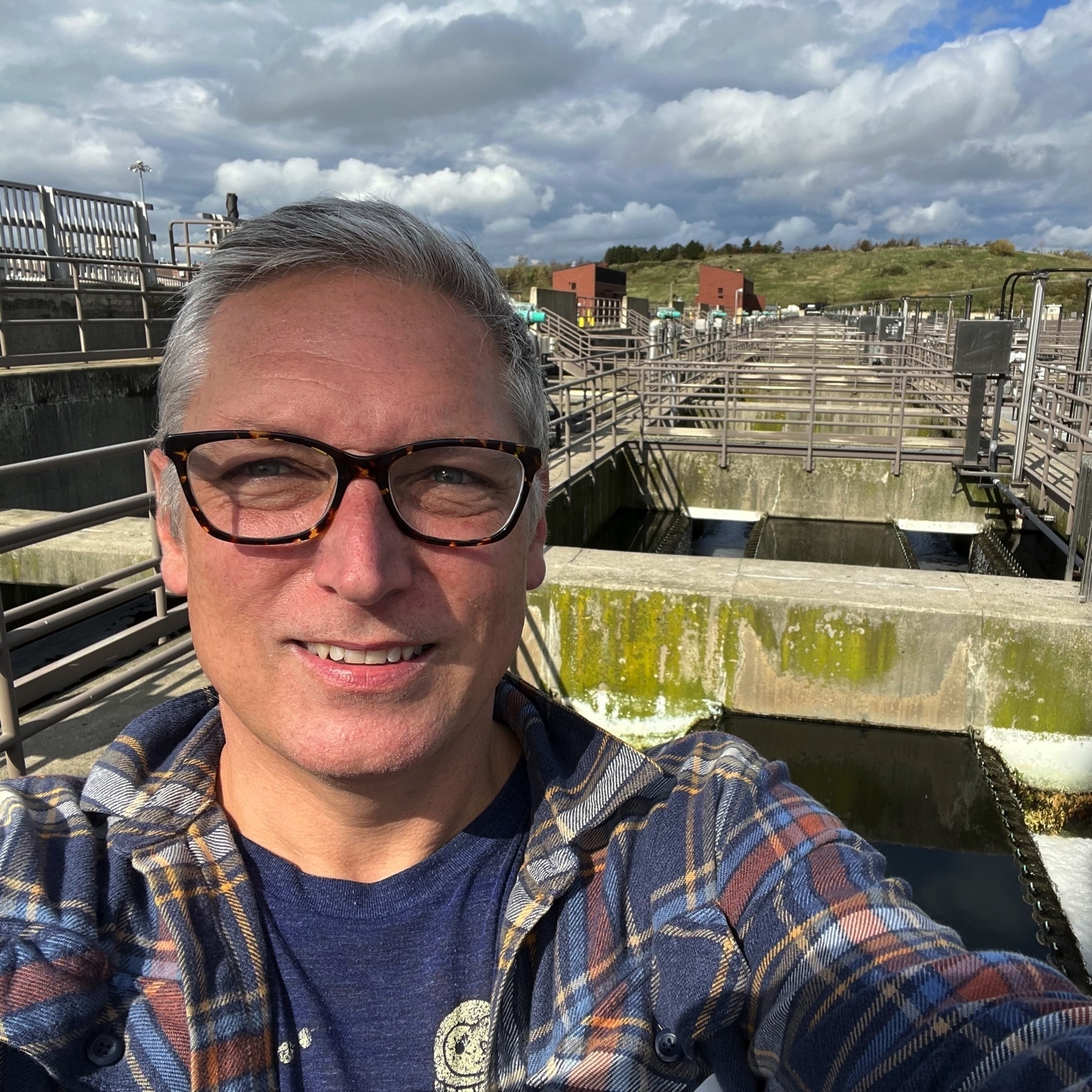 picture of myself squinting into the sun, with grey hear, reddish skin, and a blue plaid shirt. expansive water treatment structures behind me.