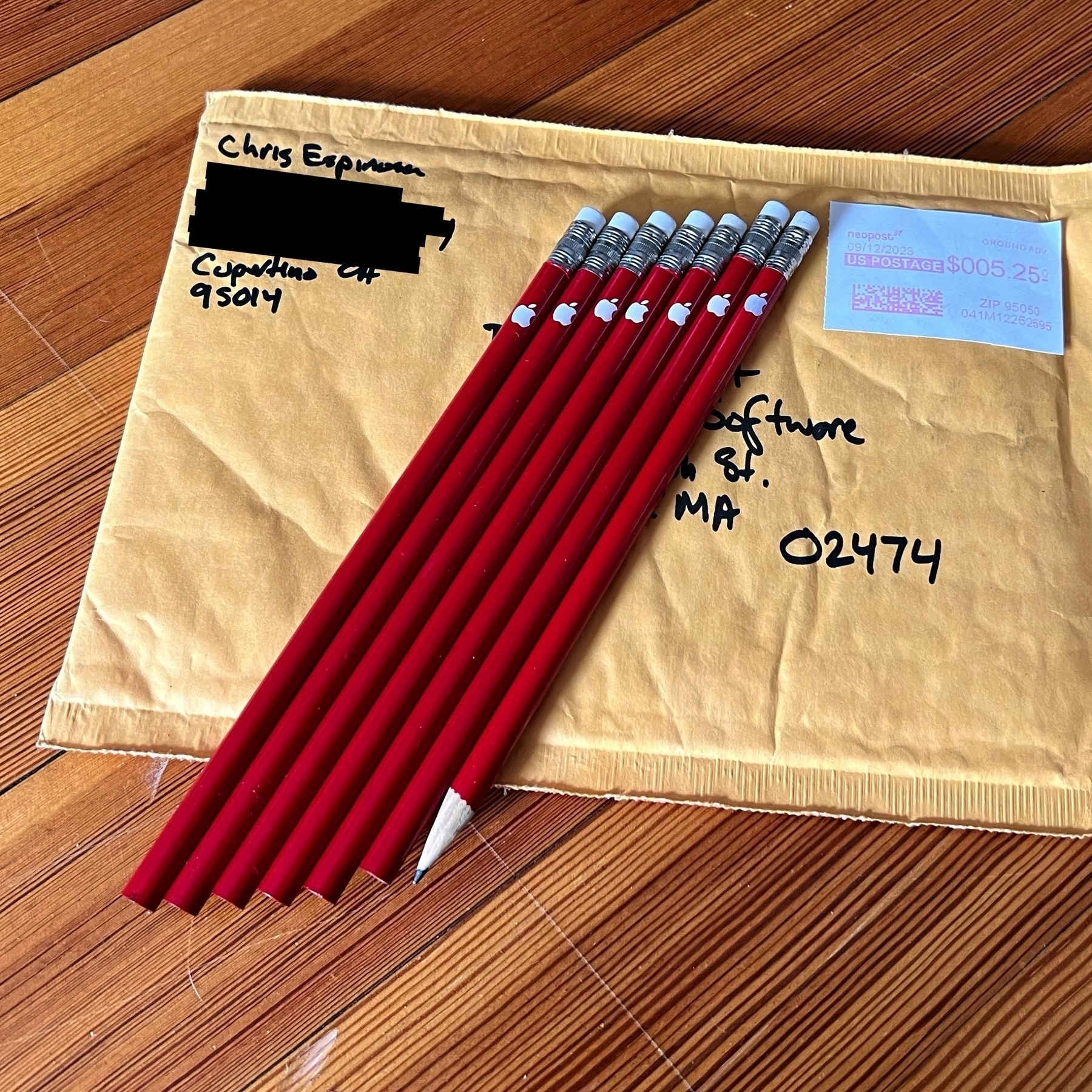 Picture of seven red pencils with white Apple logos, arranged in a diagonal row on top of a shipping envelope