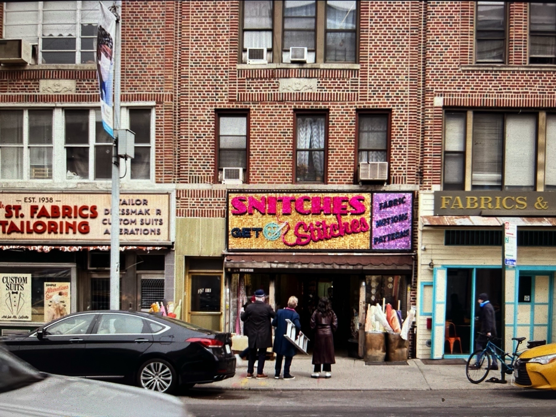 Still capture of a scene from the Hulu show “Only Murders in the Building”, showing a New York streetfront with a business called “Snitches Get Stitches”