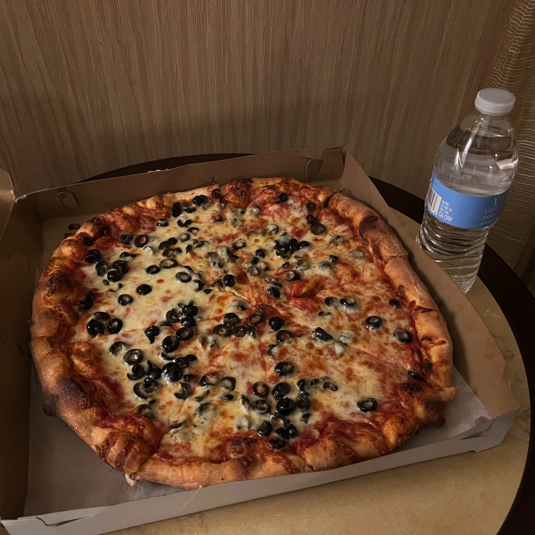 Picture of a cheese and black olive pizza in a box, next to a small water bottle. The pizza looks large for a small pizza.