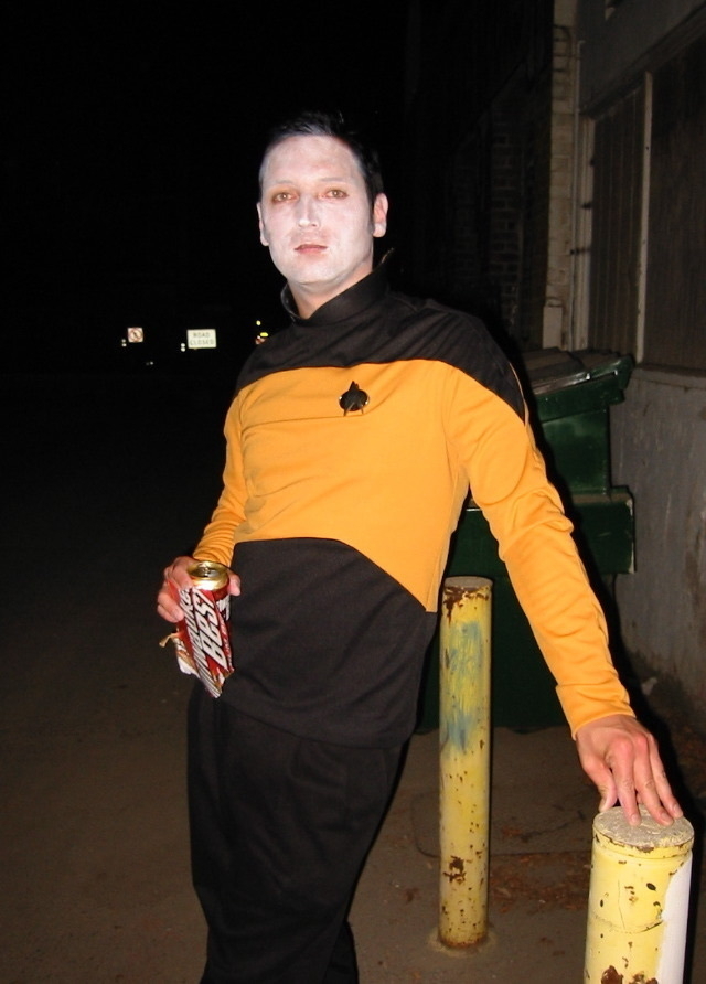 Man with black hair and a star trek uniform, holding a beer, with white make-up making him look more like Data from Star Trek.