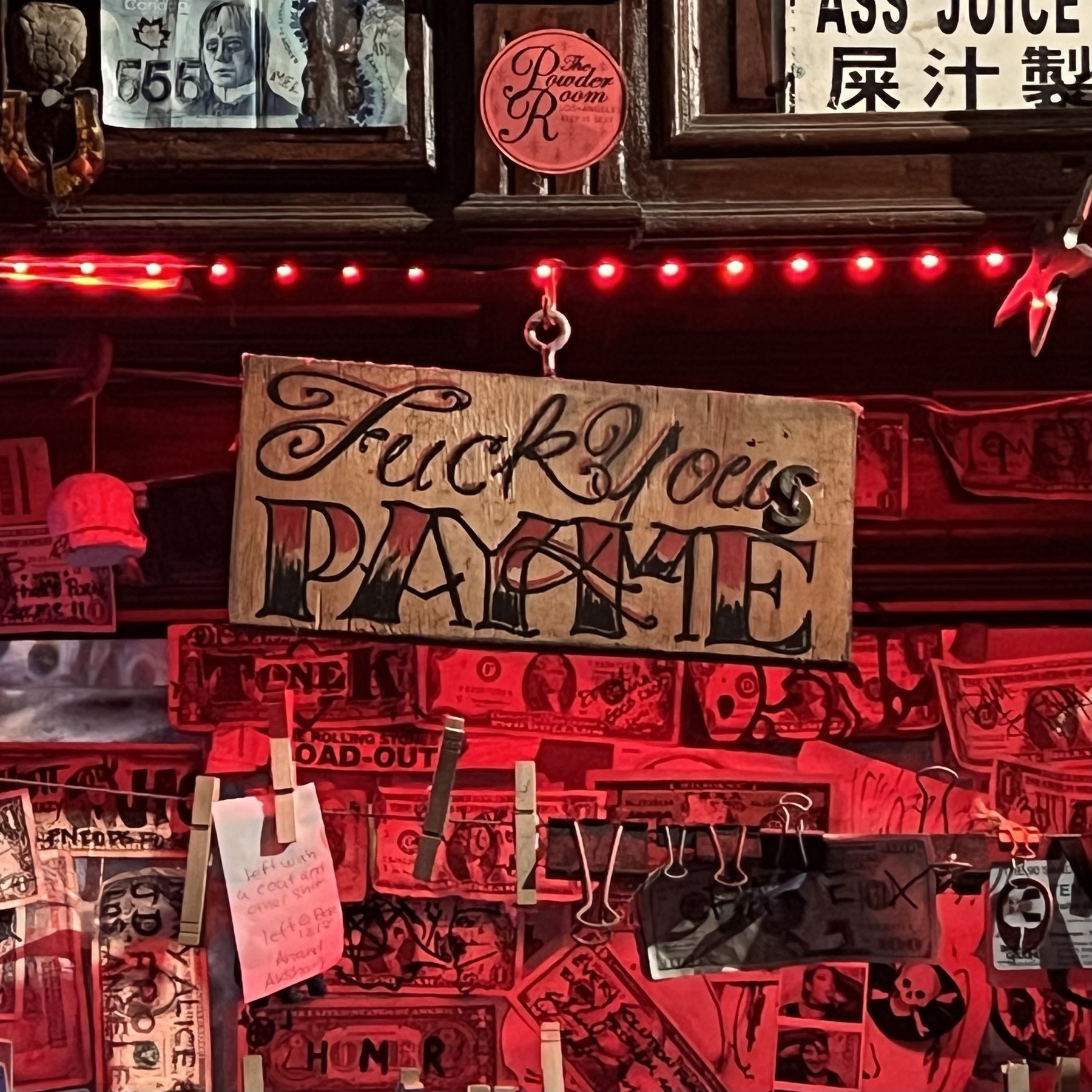 Photo of a sign hanging behind a bar among colorful red lights and autographed dollar bills. The sign reads “Fuck You(s) Pay Me”