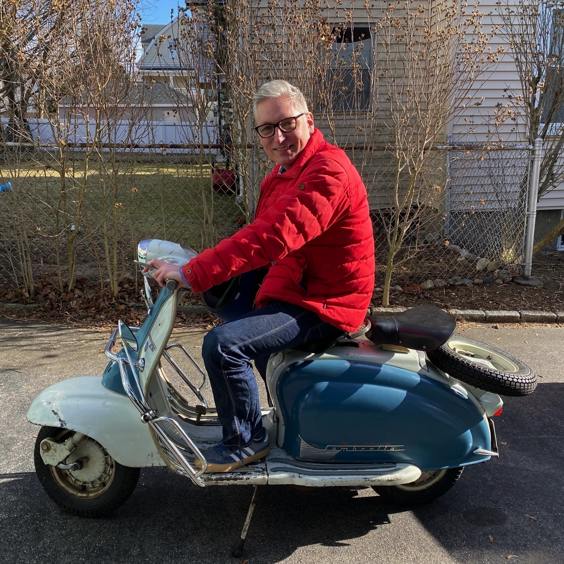 Caucasian man with grey hair wearing a red, puffy jack, sitting on a classic Lambretta motorcycle with blue and white trim.
