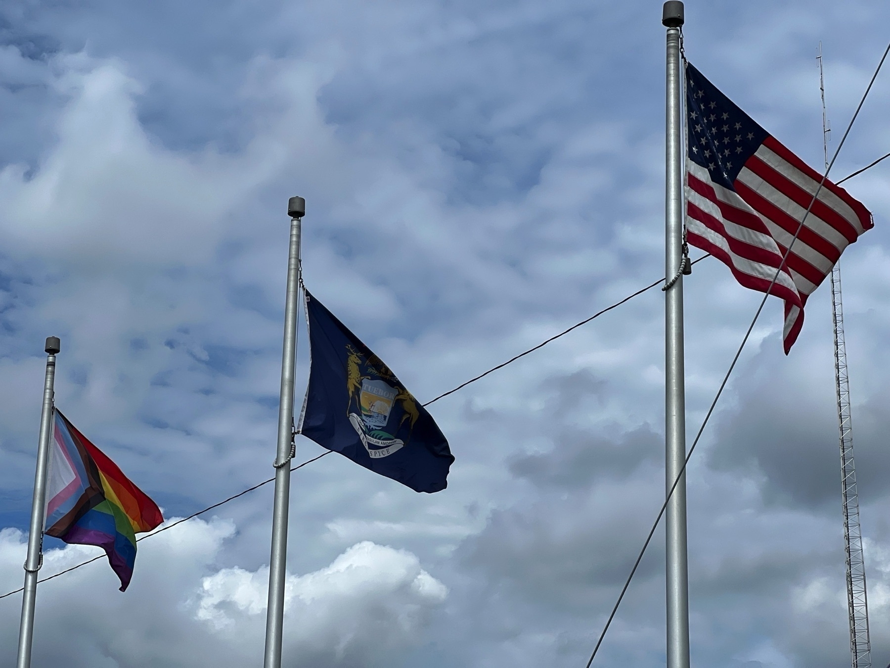 Three flagpoles against a partially clouded sky: one US Flag, one Michigan Flag, and one Pride Flag.