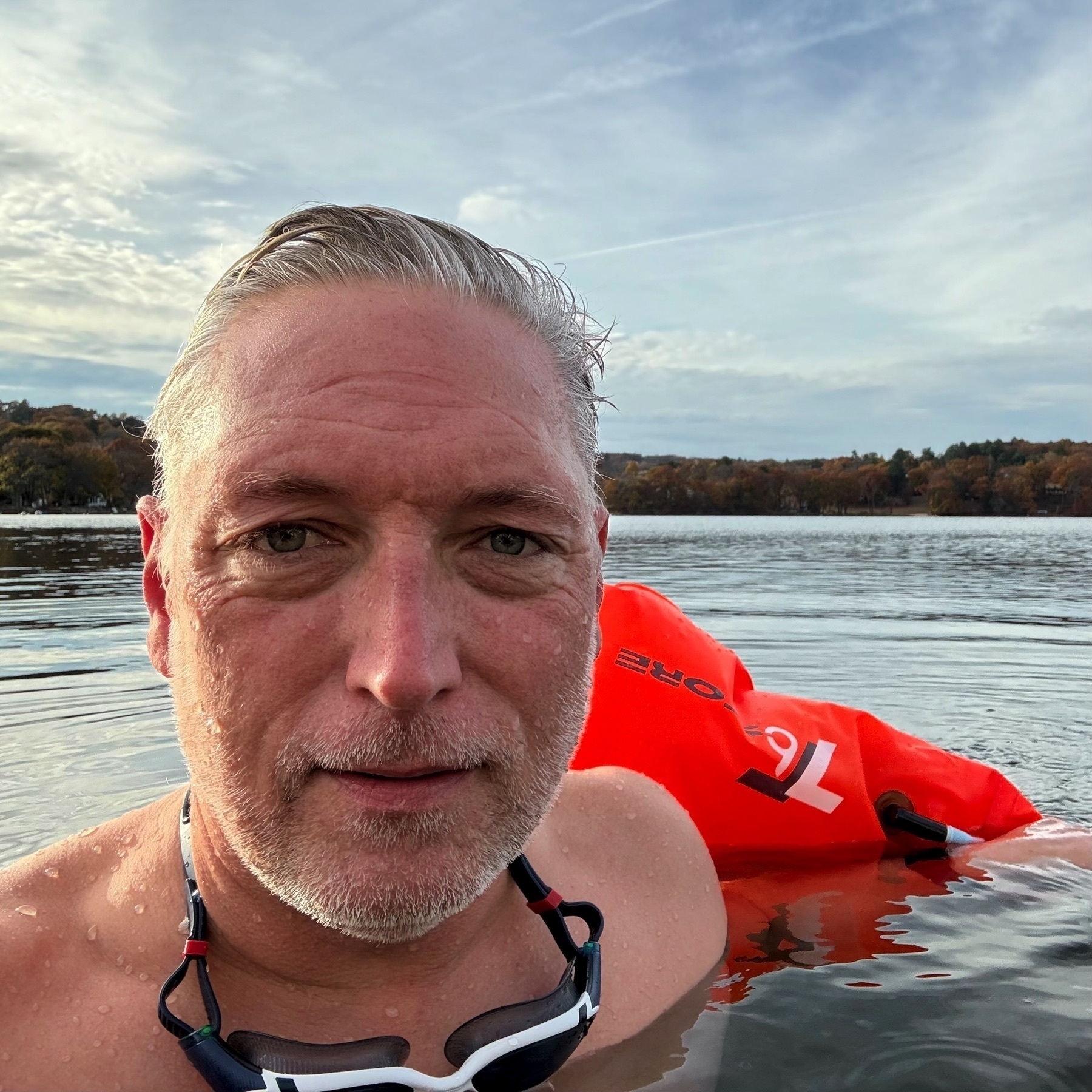 Caucasian man, chest high in lake water, looking into the camera. Backdrop of calm water with trees on the opposite shore.