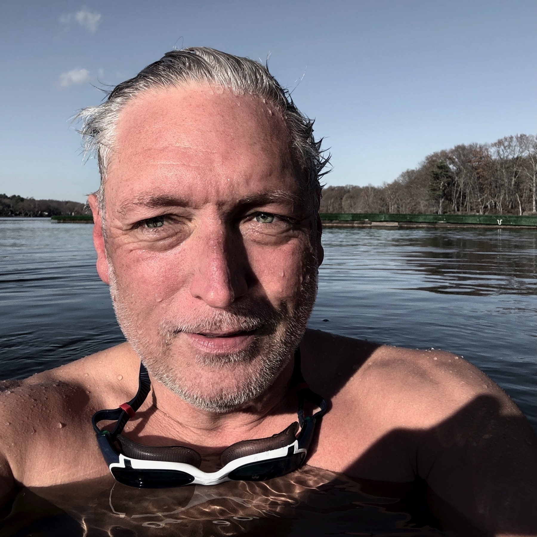 Selfie of a man shoulder-high in lake water with a backdrop of sky and trees.