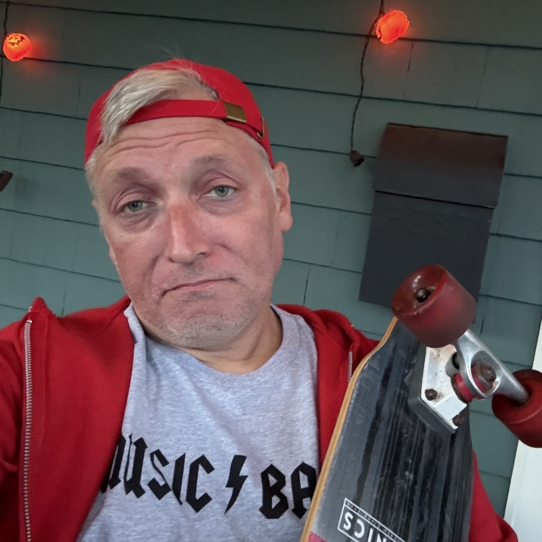 White man in costume as Steve Buscemi’s character from 30 Rock, wearing a red sweatshirt, grey “Music Band” t-shirt, and red baseball cap on backwards. Face has sunken eyes, looking into the camera with a smug expression.