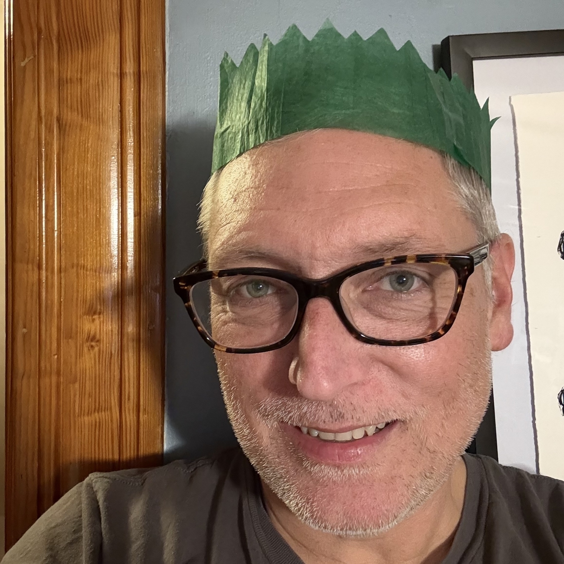 Caucasian man wearing glasses, with white facial hair stubble, smiling, staring into the camera and wearing a green paper crown.