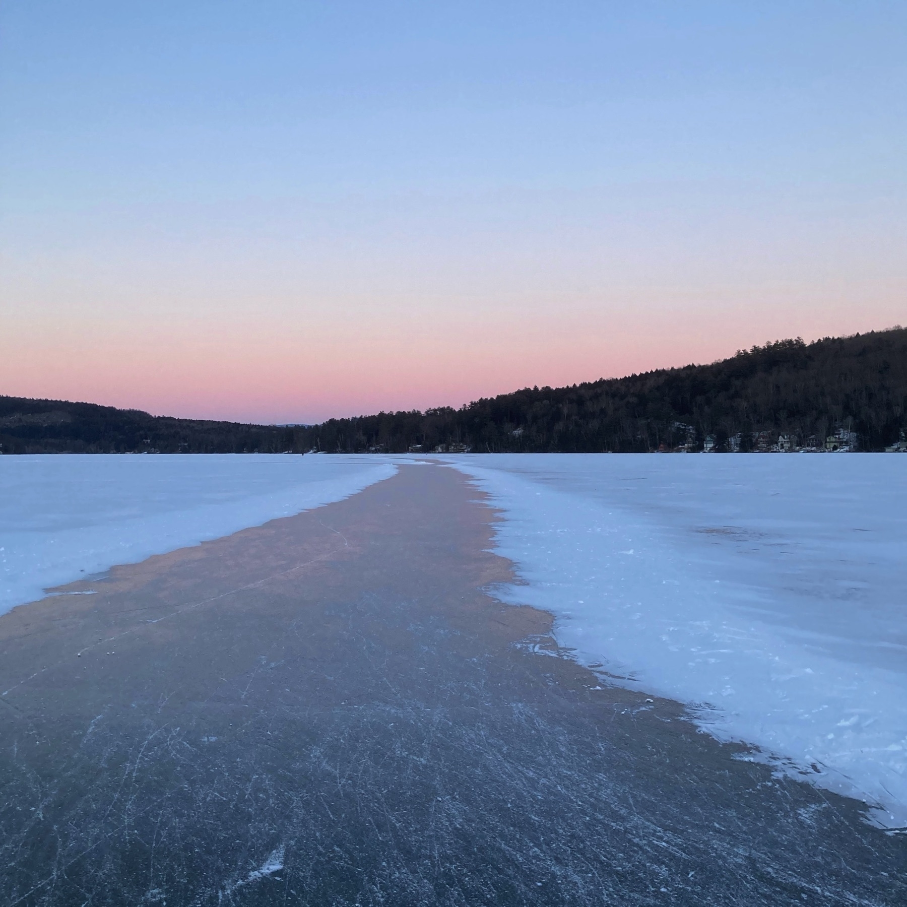 Frozen lake with snow covering everything except a path of ice with ice skate scuff marks. Sunset behind hills in the distance.