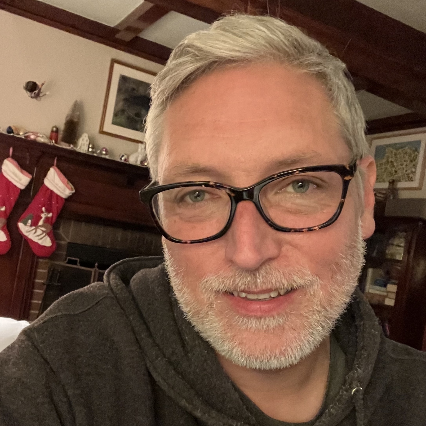 Selfie of a man with gray hair, a reddish complexion, wearing glasses, and gray/white beard, about 1/4 inch long. Background a living room with stockings on a fireplace mantel.