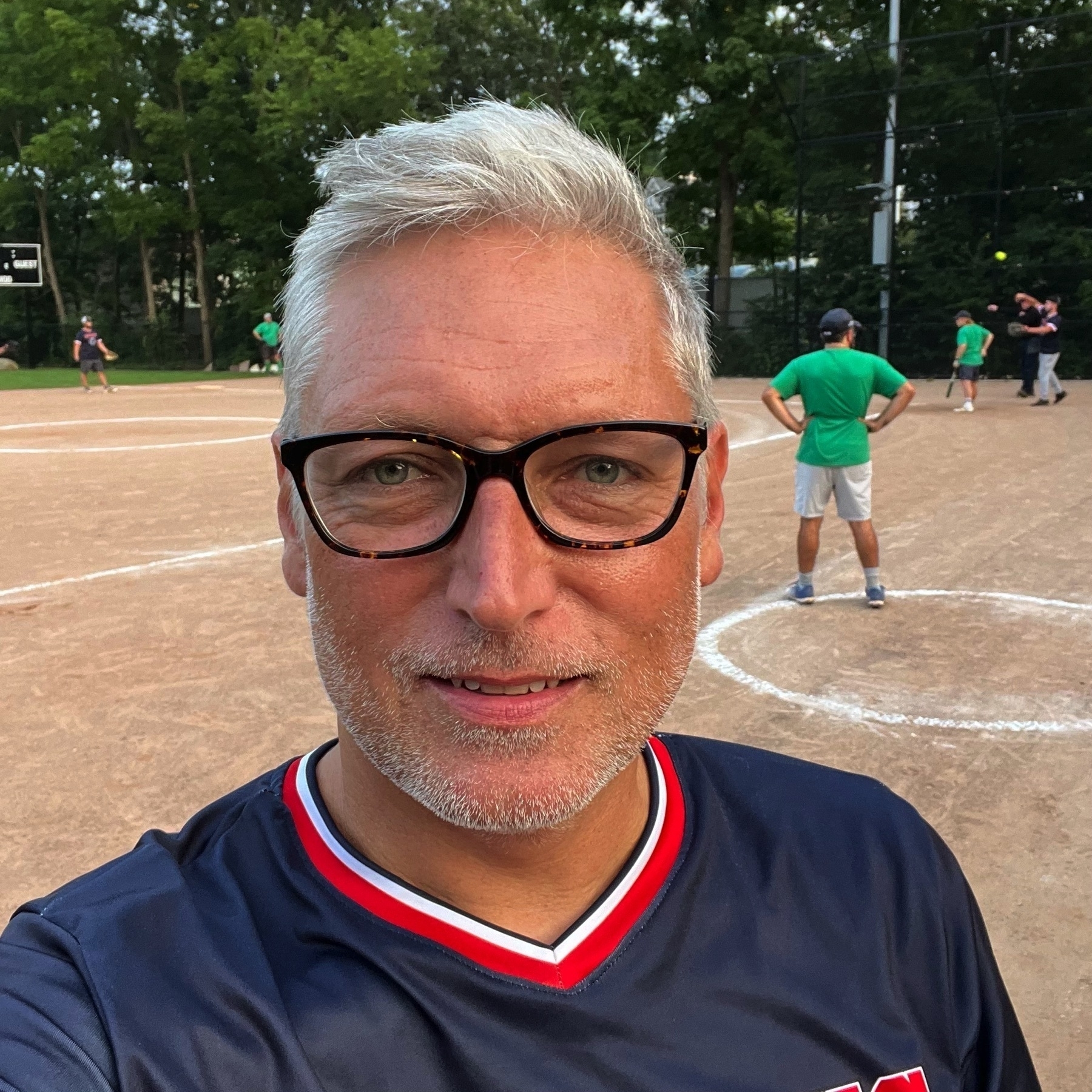 Selfie of a man with tan caucasian skin, a white stubble beard, and white/grey hair. Wearing glasses, wearing a blue baseball jersey and looking right at the camera. Baseball diamond in the background.