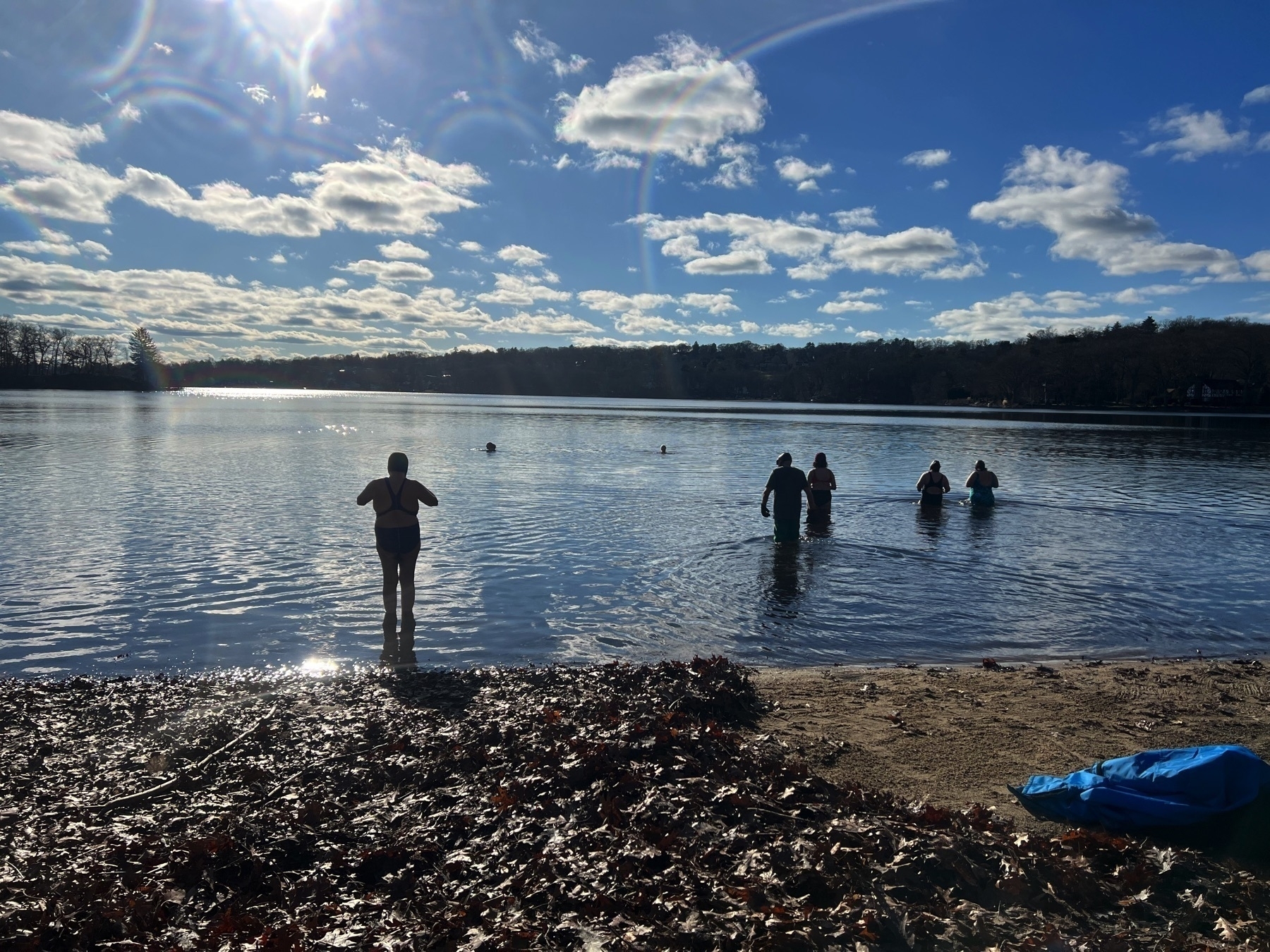 Large lake with leaf-covered beach, several people wading into the water. Blue sky with clouds.