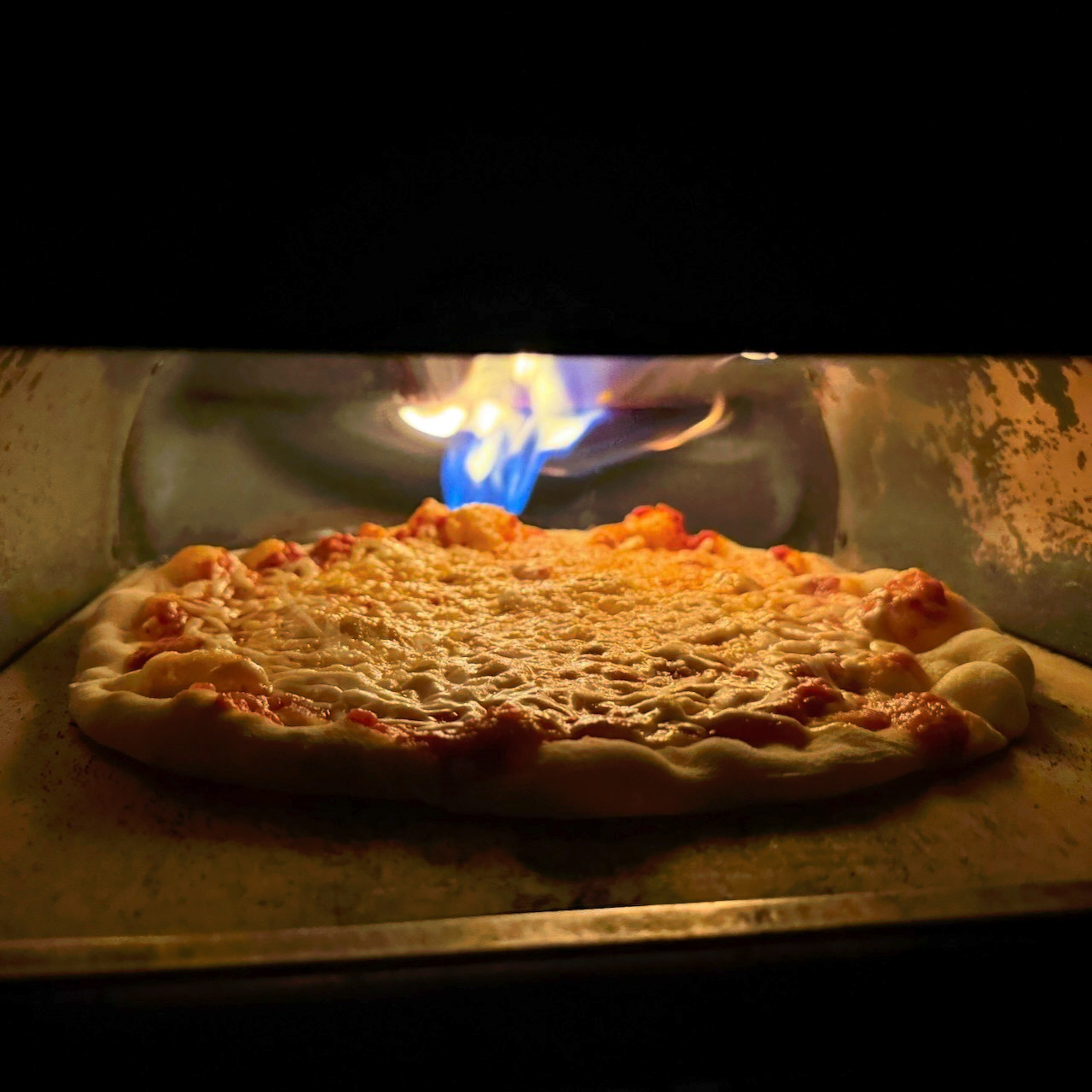 A pizza baking in the oven with a bright flame at the back of the oven, illuminating a pizza with boiling cheese and sauce under the intense heat.