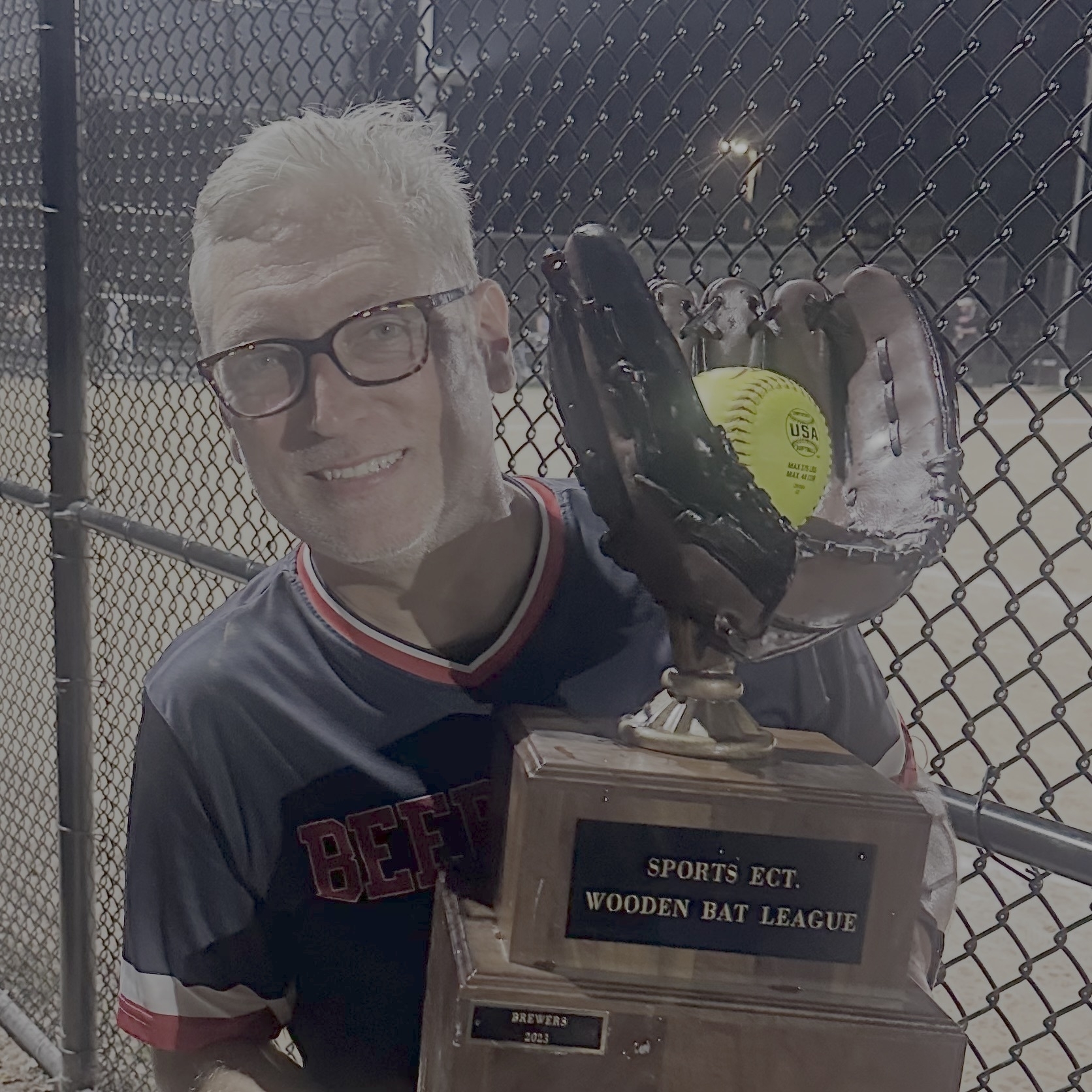 Selfie of a caucasian man with white hair, glasses, smiling while holding a trophy in the shape of a softball mitt with a yellow ball in it.