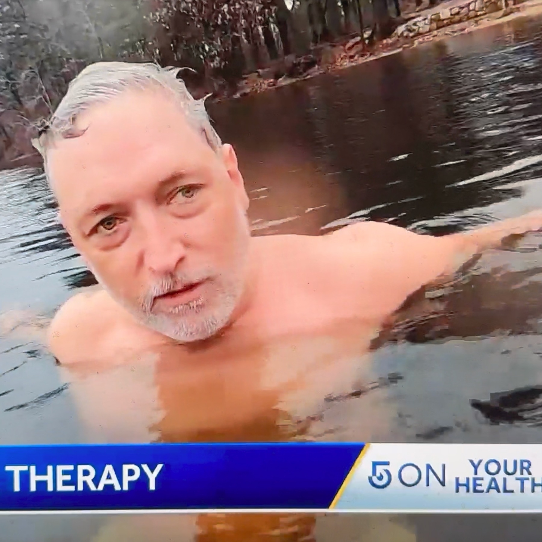 Still from a local news broadcast showing a caucasian man with grey hair (Daniel) wading neck-high through lake water.