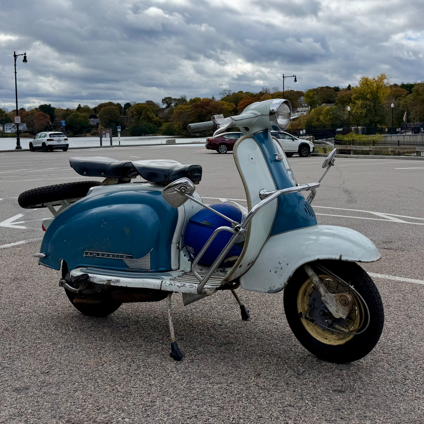 1962 blue and white Lambretta scooter in a parking lot, lake and treeline, with blue sky and clouds in the rear.