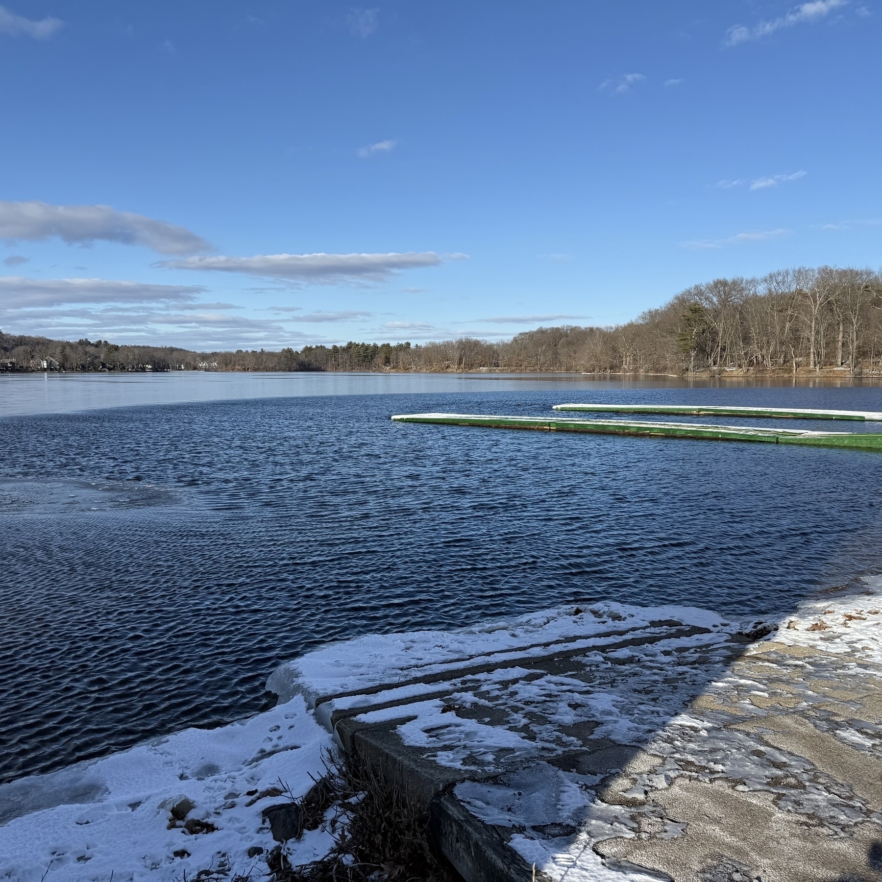 Iced over lake with open water surrounding two snow dappled docks. Clear skies and bare winter trees on the other side of the lake.