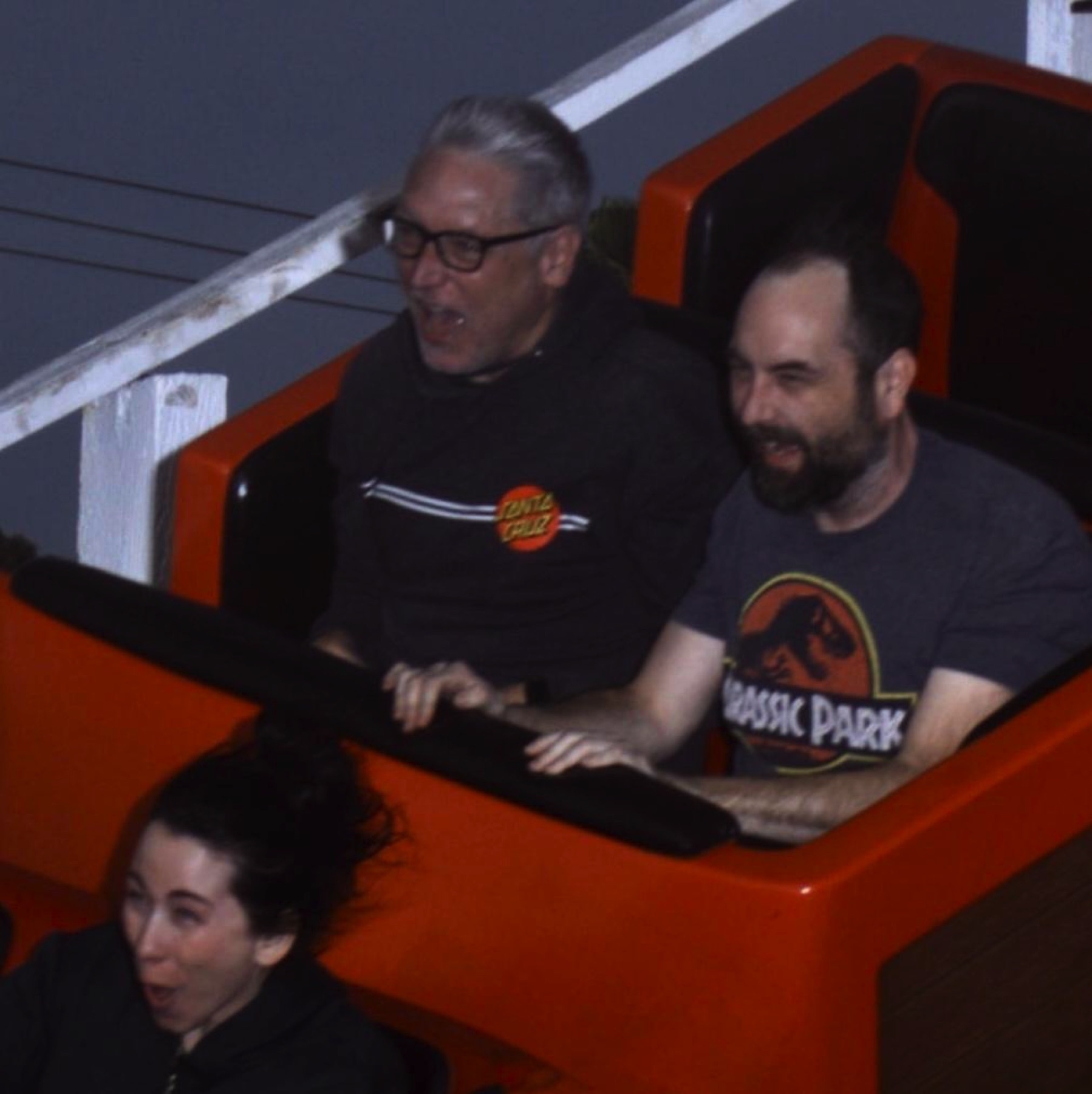 Picture of two men seated together in a roller coaster carriage, mouths open and hair swept back in the thrill of the ride. A woman in front of them is visible with a similar expression.