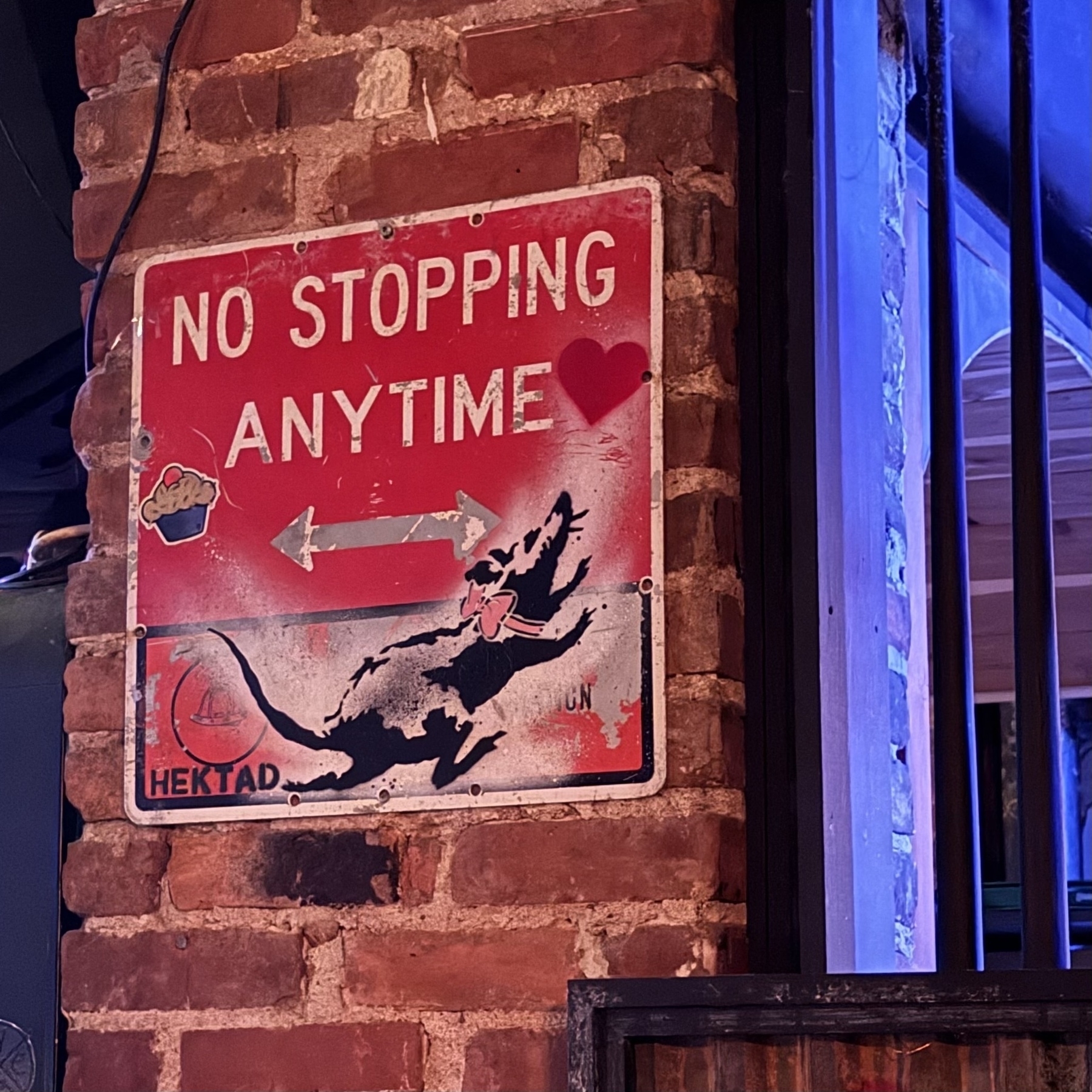“No Stopping Anytime” street sign mounted on a brick interior wall, next to a window with security bars. The sign is adorned with a Banksy-style stencil of a rat.