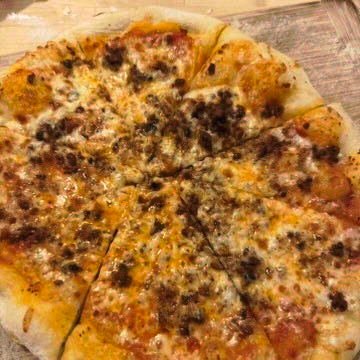 Top-down picture of a home-made pizza on a cutting board. Specks of semolina all around the pizza. Pizza is cut into 8 with a charred look and many brown, meat-like chunks on top of the cheese.