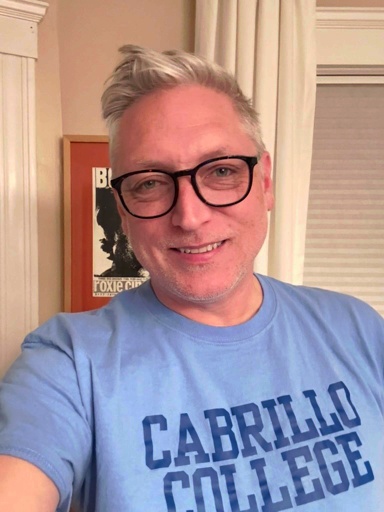 Picture of a caucasian man wearing black framed glasses, looking into the camera, smiling. He's wearing a blue t-shirt with typical squared font reading "CABRILLO COLLEGE".