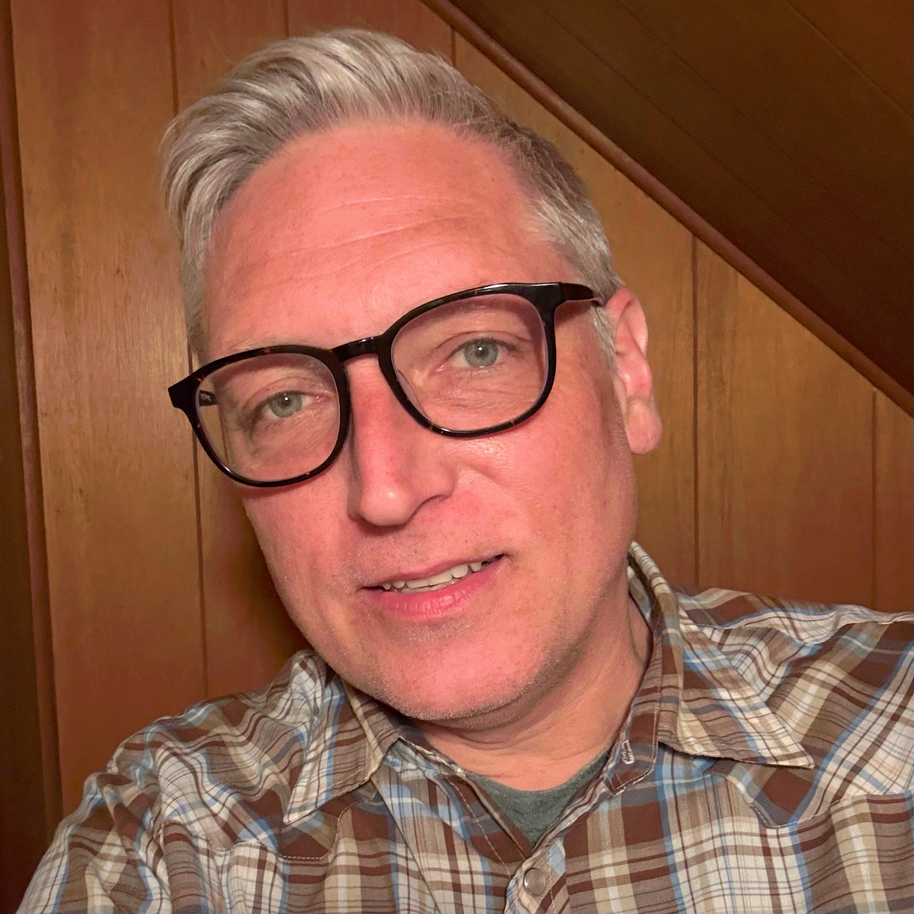 Selfie of a 50-year-old caucasian man with short grey hair and glasses, wearing a brown, blue and white plaid “western” shirt.