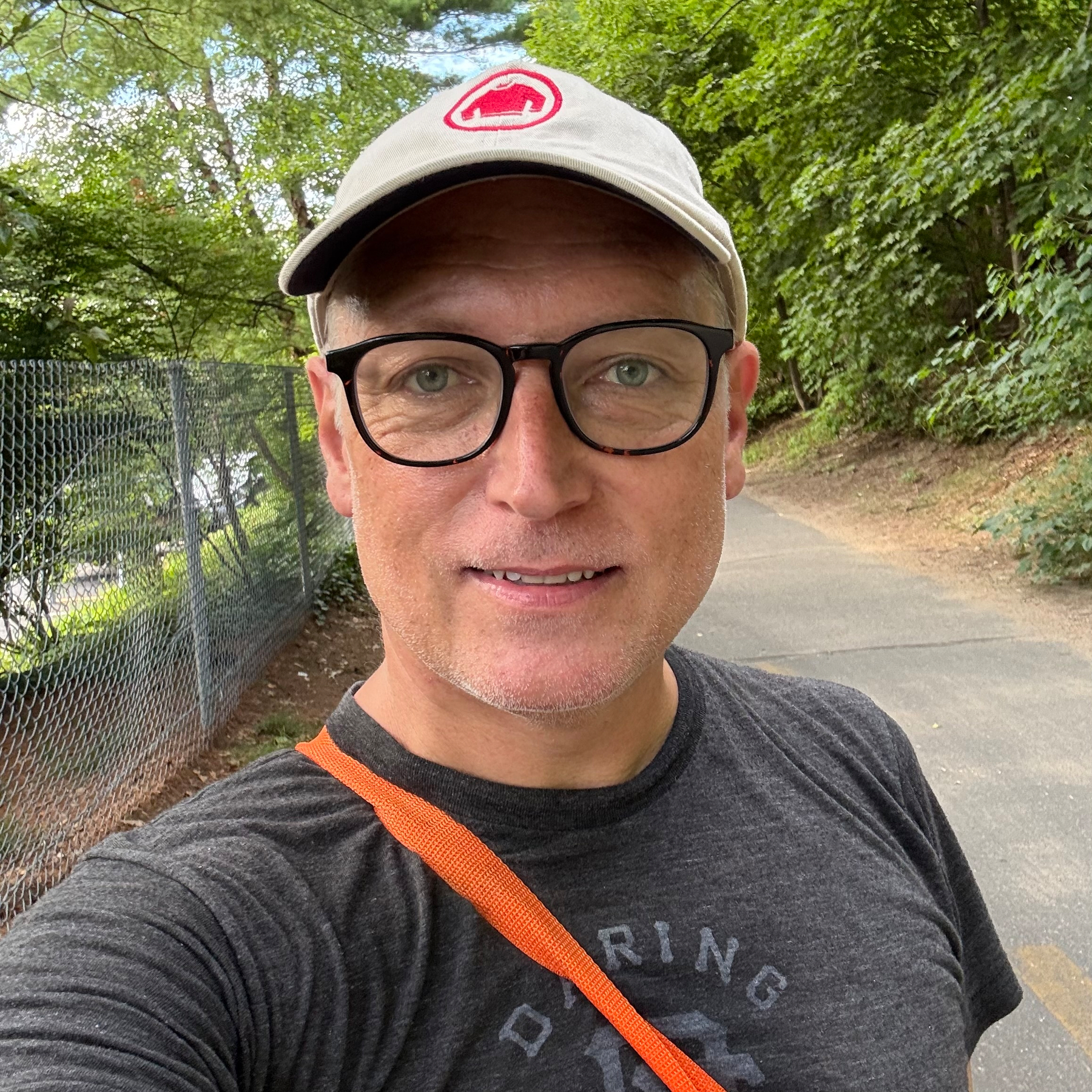 Picture of a white man with tan face and dark glasses, looking directly into the camera. He's wearing a beige cap with a "Red Sweater" logo, and heathered black t-shirt with "Daring" visible. An orange strap across his chest. Background is a bicycle path with trees alongside.