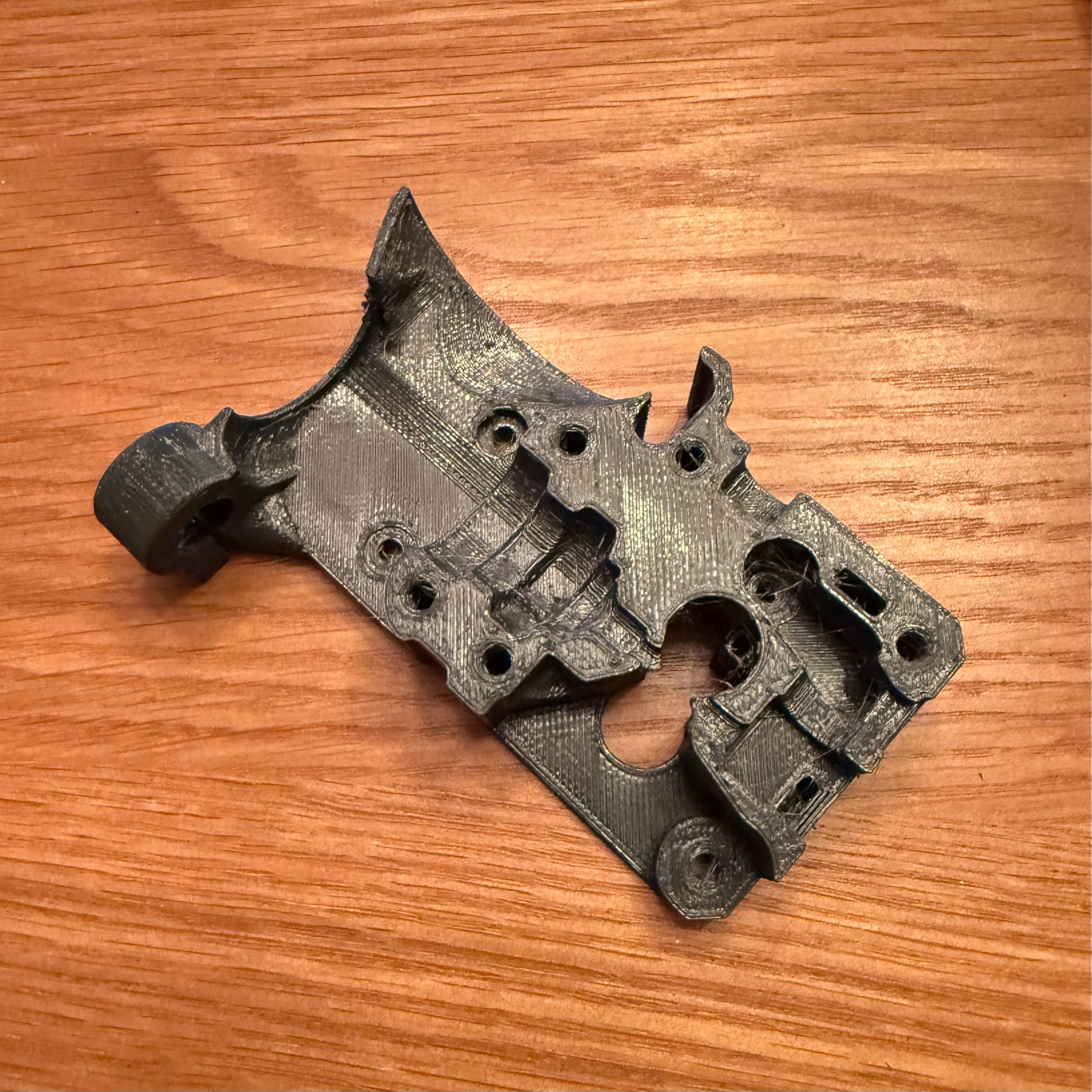 3D-printed component that serves as part of the Prusa MK3 line of printers. Material is a dark blue filament against a wood table. Otherwise the contours are probably not interesting.
