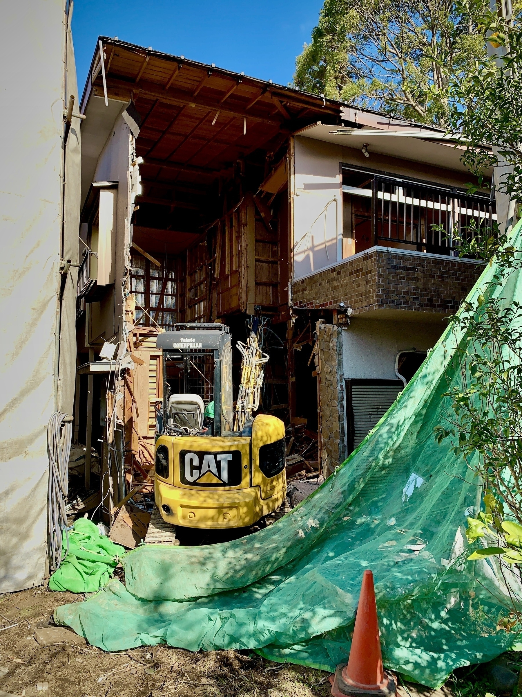 A home between white and green tarps being torn down by a small yellow backhoe. 