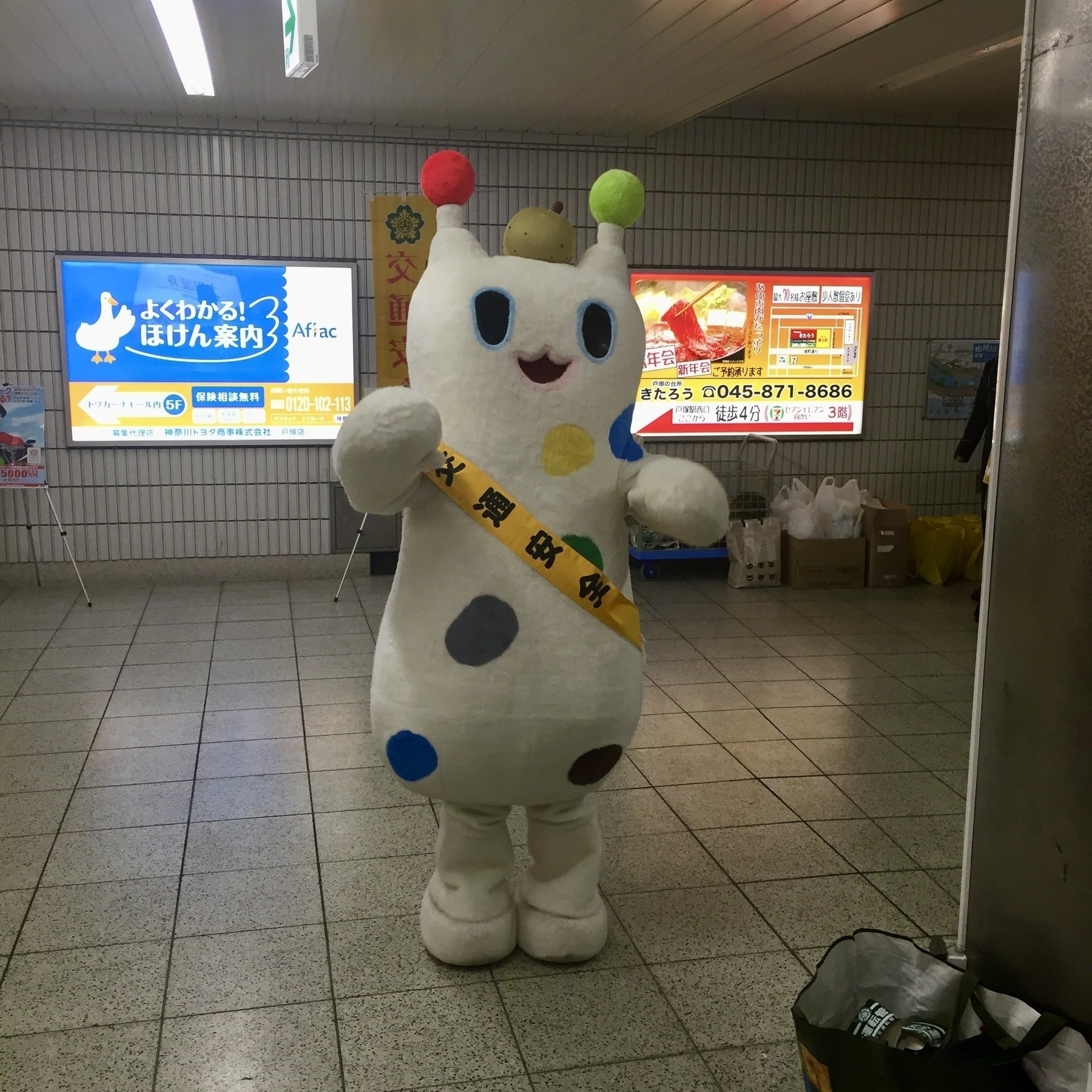 A person in a mascot suit, with polka dots in green, blue, and yellow, and antennae, has a sash with “koutsu anzen” (traffic safety) written on it. 