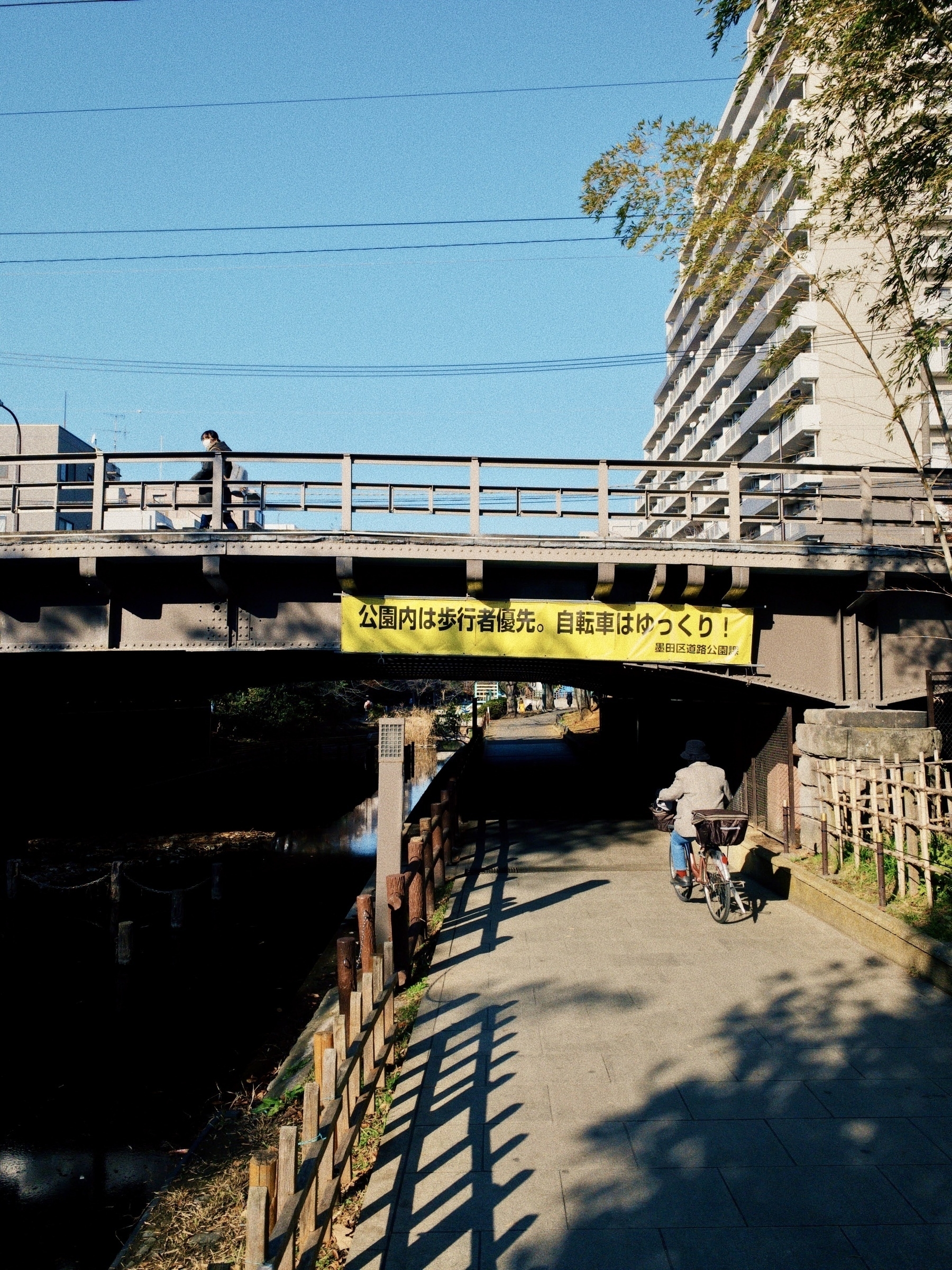 A bike rider in a grey jacket rides under an underpass while a walker crosses the bridge above. The yellow sign on the bridge admonishes riders that pedestrians have right of way, and to ride slowly. 