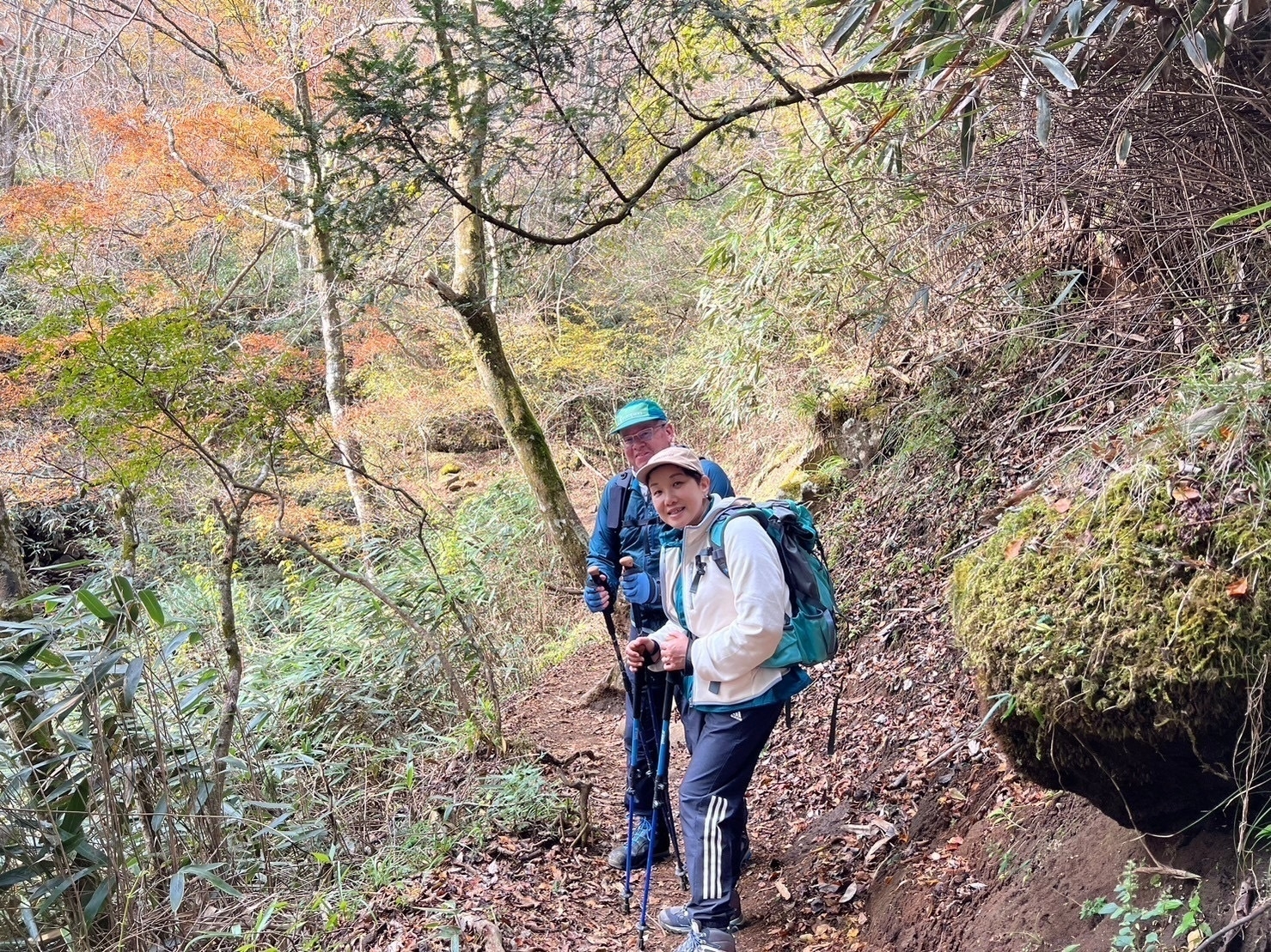 Woman in white and man in blue hold hiking poles and wear backpacks on a hiking trail. 