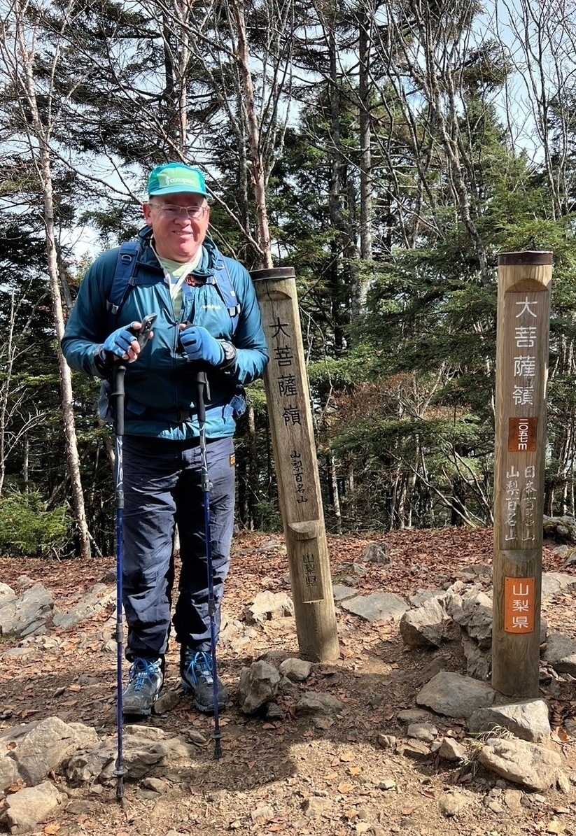 Male hiker at summit of Mt. Daibosatsurei in Yamanashi Japan, wearing black pants and a teal colored jacket, holding hiking poles next to the summit marker. 
